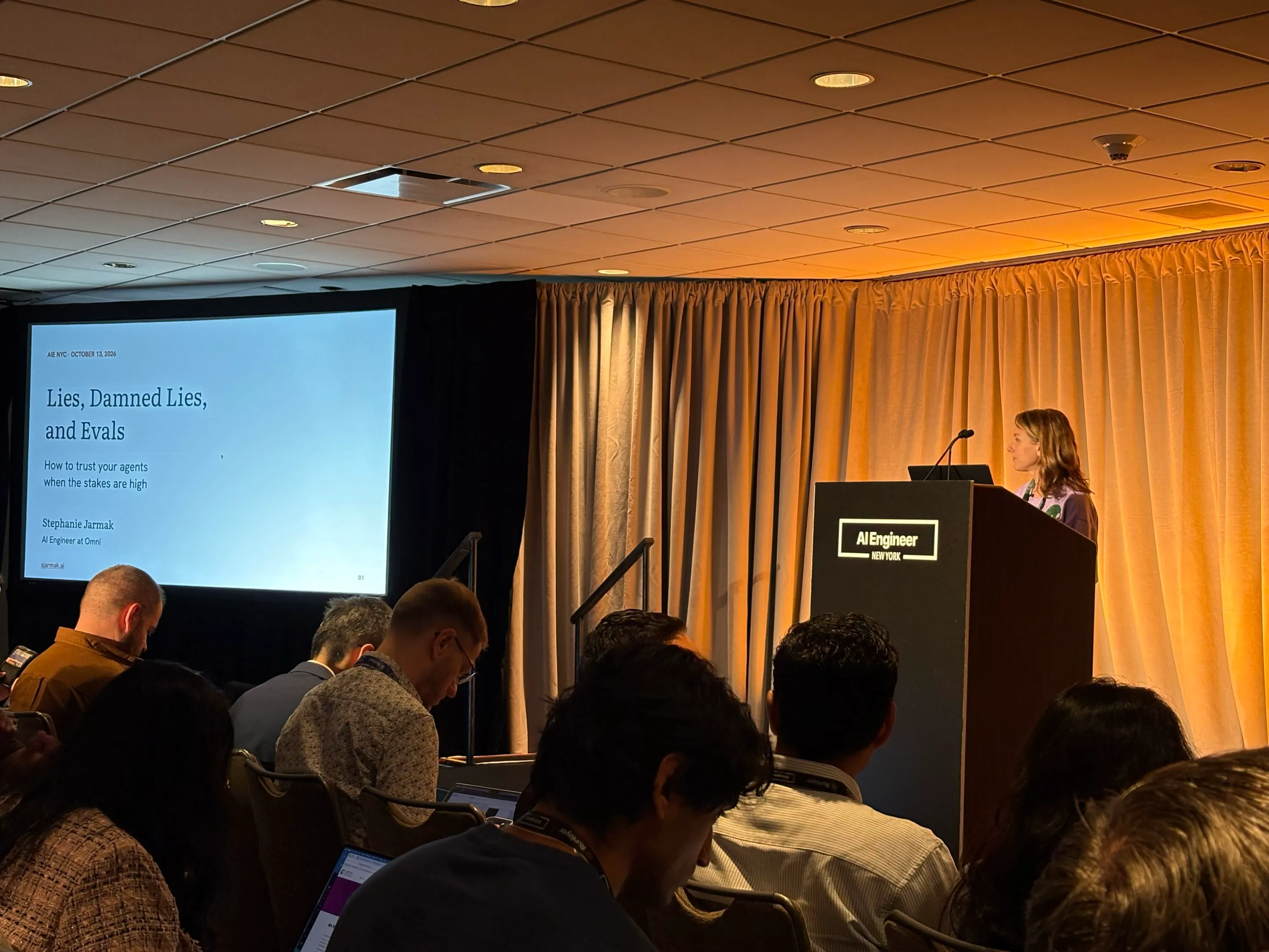 Stephanie Jarmak at the podium at AI Engineer New York, October 13, 2026, with the title slide "Lies, Damned Lies, and Evals: How to trust your agents when the stakes are high" on screen and the audience seated in front.